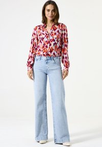 Blusa colorida y estampada con mangas largas y cuello, combinada con unos vaqueros de pierna ancha en azul claro y zapatos color blanco roto. Textura de tela suave.