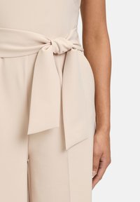 Tuta beige con una cintura ampia legata. Il materiale appare liscio, con una leggera lucentezza, ed è sagomato per una silhouette aderente.