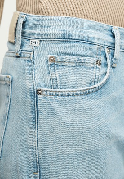 Φωτεινό μπλε τζιν τζιν με μπροστινή τσέπη με μεταλλικά καρφιά και ετικέτα CofH, φορεμένο με μπεζ ριμπ πλεκτό τοπ.