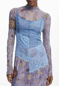Vestido de mezclilla con silueta ajustada, que presenta un overlay de encaje azul y mangas de malla con patrón floral púrpura. Tejido texturizado y detalles sutiles.