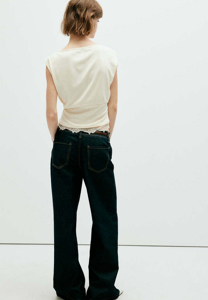 Femme aux cheveux courts portant un haut blanc sans manches avec une bordure en dentelle et un jean large foncé, debout, mains dans les poches, de dos.