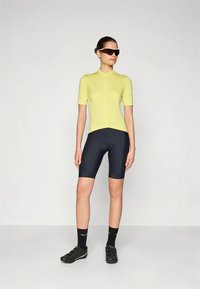 Tenue de cyclisme composée d'un haut jaune vif à manches courtes et d'un short bleu foncé. Le mannequin porte des lunettes de soleil et des chaussures de cyclisme noires.