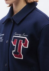 Tamsiai mėlynas vilnos švarkas su užsegamu priekiu, dideliu raudonos ir baltos spalvos siuvinėtu logotipu bei kontrastingu baltu siuvimu. Apykaklės dizainas klasikinis.