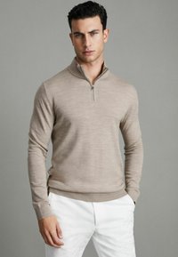 Pulover bej cu fermoar, cu mâneci lungi, confecționat dintr-un material moale. Prezintă un guler înalt și manșete ribbate. Este purtat cu pantaloni albi.