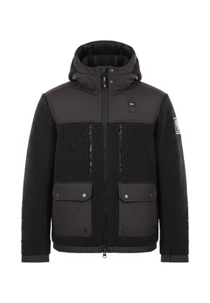 Giacca nera a doppia texture con parte superiore liscia e sezione inferiore pelosa, tasche con zip, cappuccio regolabile e patch con logo sulla manica.
