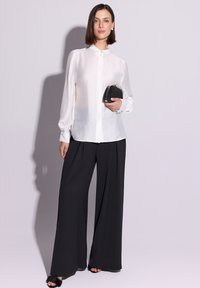 Przezroczysta biała koszula z długim rękawem zapinana na guziki, zestawiona z szerokimi czarnymi spodniami. Trzyma małą czarną kopertówkę. Prosty, elegancki design.
