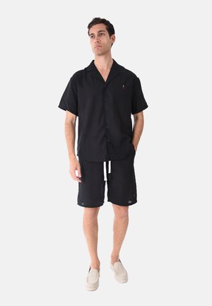 Uomo che indossa una camicia nera a maniche corte con bottoni e pantaloncini neri con coulisse, con scarpe beige slip-on, in piedi davanti a uno sfondo neutro.