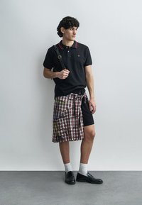 Tricou polo negru cu accente roșii, fustă wrap cu model în carouri purtată peste pantaloni scurți negri, șosete albe și pantofi negri, pe un fundal minimalist.