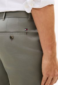 Γκρι παντελόνι με λεία υφή, που έχει τσέπη πίσω και καφέ κουμπί, με μια μικρή λογότυπο σε κόκκινο, λευκό και μπλε χρώμα.