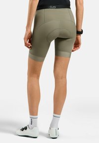 Olijfgroene fietsbroek met platte naden, een naadloze gepolsterde zone en een brede tailleband, gecombineerd met witte fietsschoenen en sokken.