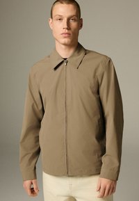 Chaqueta marrón claro con cierre de cremallera en la parte delantera, cuello puntiagudo y mangas largas. Hecha de un material suave y ligero sin patrones visibles.