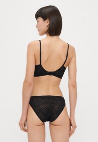 Črn komplet spodnjega perila vsebuje gladko braletko z nastavljivimi naramnicami in ujemajoče se nizko pasne hlačke, oboje okrašeno z diskretno listnato vzorcem.