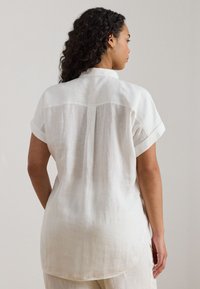 Balta linu krekls ar apkakli, īsām piedurknēm un uzlocītām aprocēm. Ar centrālu šuvi muguras daļā un nedaudz izliekta apakšdali. Teksturēts audums.