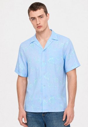 Jeune homme portant une chemise à manches courtes bleu clair avec un motif subtil de tortue et un jean bleu sur un fond uni.