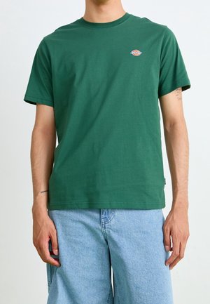 Mand iført en grøn kortærmet t-shirt med et lille logo på brystet og lyseblå denimshorts.