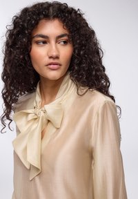 Blusa di seta color crema con un grande fiocco al collo, caratterizzata da una texture liscia e morbida e una chiusura con bottone al collo.