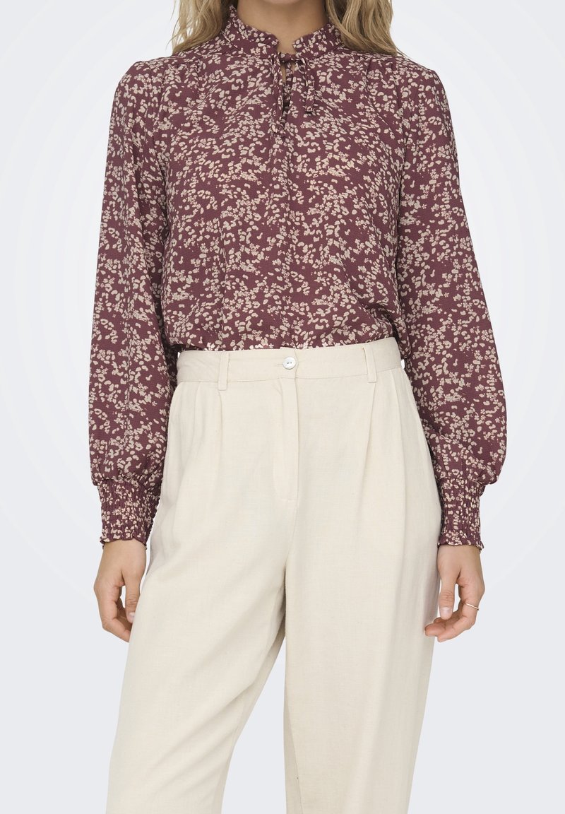 Femme portant un chemisier bordeaux à manches longues avec un petit motif floral et un pantalon large taille haute couleur crème.