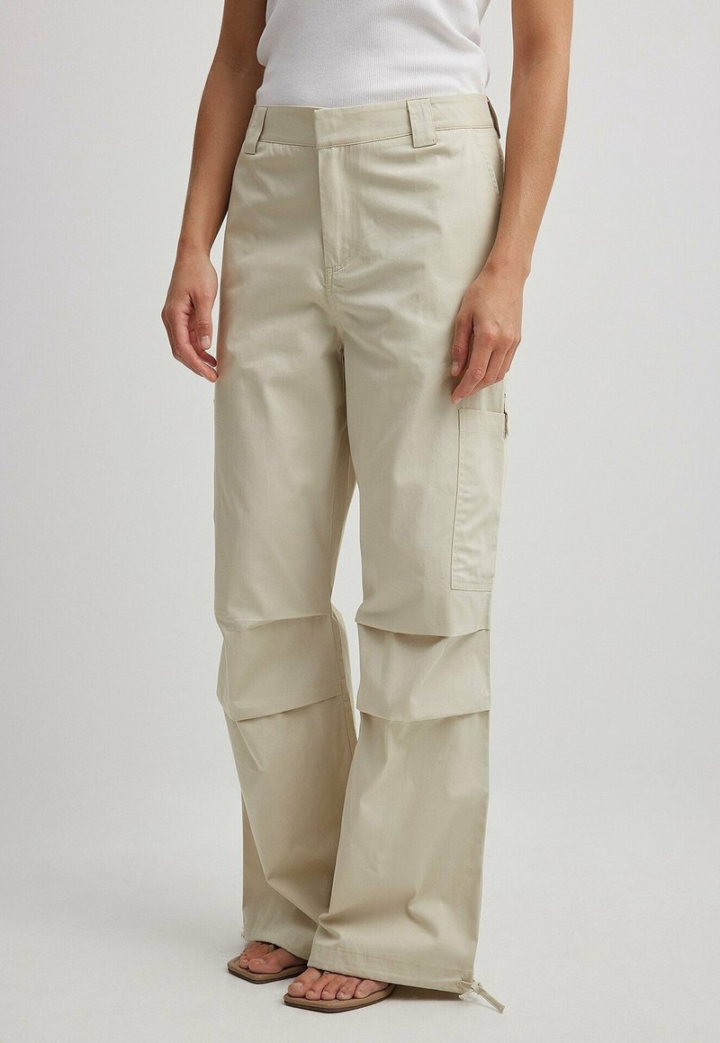 Pantalon cargo beige à jambes larges avec poches latérales et détail zippé à l'ourlet, porté avec un haut blanc sans manches et des sandales.