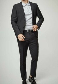 Veste noire sur un polo gris, coupe ajustée, avec un pantalon noir slim et des chaussures noires brillantes. Des boutons simples et des détails de ceinture sont visibles.