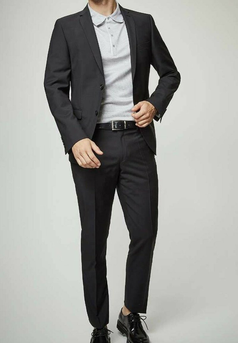 Veste noire sur un polo gris, coupe ajustée, avec un pantalon noir slim et des chaussures noires brillantes. Des boutons simples et des détails de ceinture sont visibles.