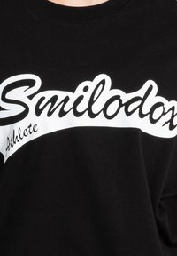 Camiseta negra con un atrevido texto blanco que dice "Smilodox" y "Athlete", con un diseño de banda curvada en el pecho.