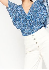 Blusa blu con motivo paisley, dotata di scollatura a V e ampie maniche a sbuffo, abbinata a pantaloni bianchi a vita alta con bottoni decorativi.