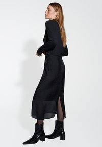 Haut noir à manches longues avec une finition texturée, associé à une jupe noire taille haute avec des fentes sur les côtés et des bottines noires en cuir brillant.