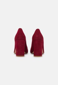 Chaussures à talons hauts en daim rouge avec un bout pointu et un large talon carré, présentant une texture lisse et un design épuré vues de dos.