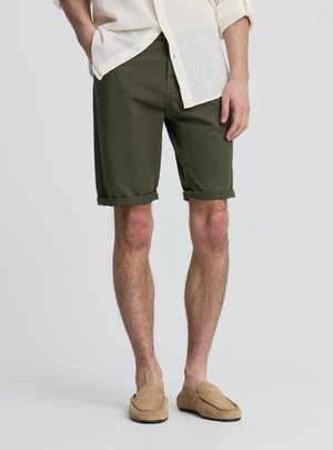 Mężczyzna w oliwkowo-zielonych podwiniętych szortach, beżowych mokasynach i kremowej koszuli z długim rękawem, stoi z jedną ręką w kieszeni.