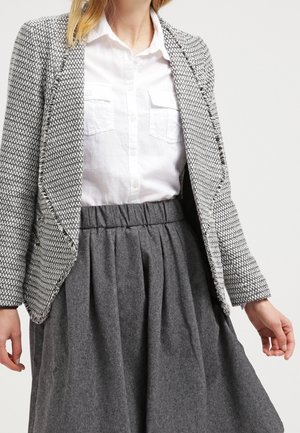 Frau trägt ein weißes Hemd mit Knöpfen, grauen strukturierten Blazer und einen hoch taillierten, plissierten grauen Rock vor schlichtem Hintergrund.