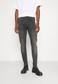 Jeans en denim gris à coupe slim avec une finition légèrement délavée, portés avec un t-shirt blanc et des bottines noires. Comprend cinq poches et une fermeture à bouton.