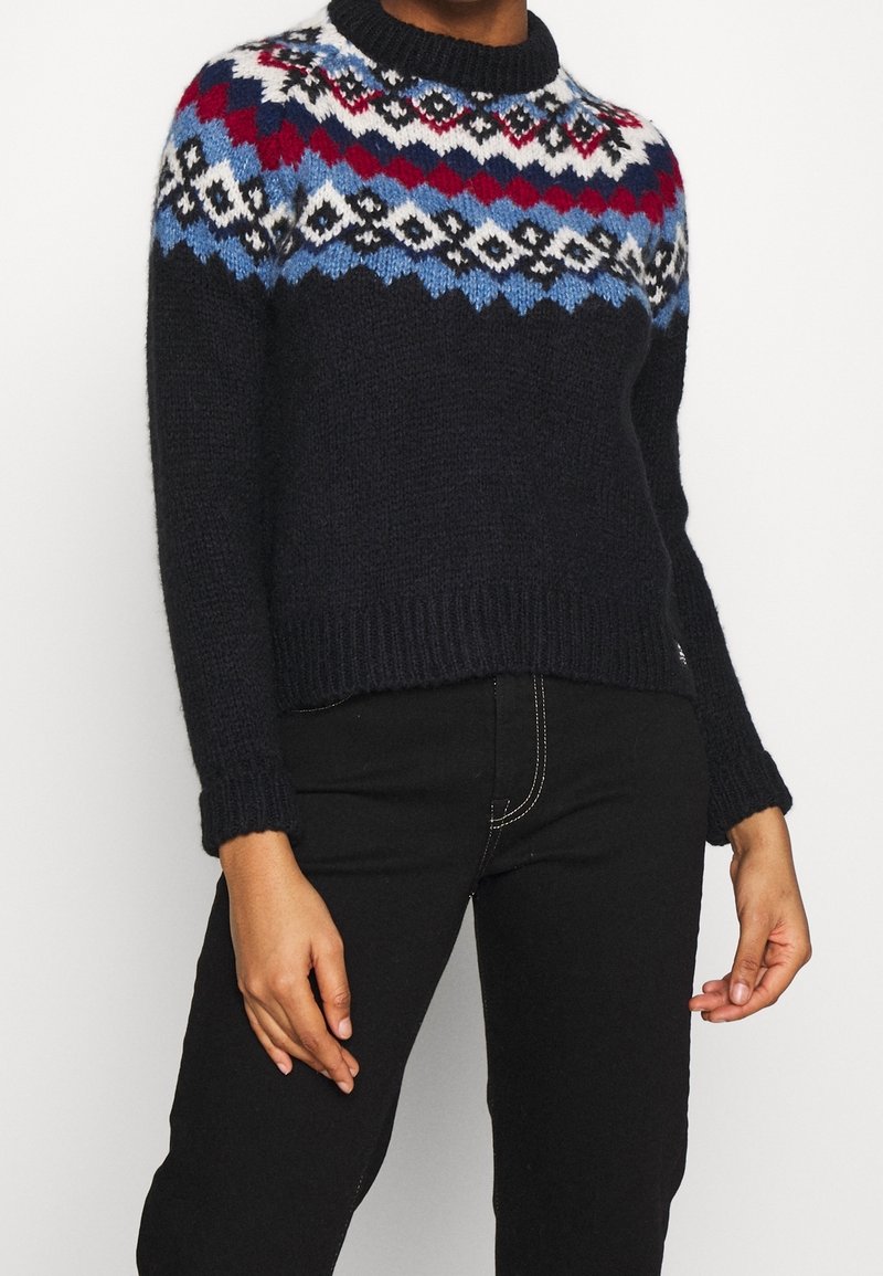 Personne portant un pull en tricot noir avec un motif géométrique rouge, bleu, blanc et noir sur la poitrine et le haut des manches, associé à un pantalon noir.