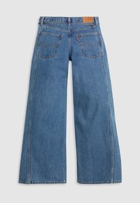 Jean en denim bleu à jambes larges, taille haute, deux poches arrière avec détails de couture, et un patch logo en cuir à la ceinture.
