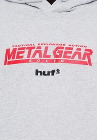 Γκρι φούτερ με μεγάλο κόκκινο λογότυπο «METAL GEAR SOLID» και μαύρο κείμενο «huf» από κάτω. Μαλακή υφή με  κουκούλα που έχει κορδόνι.