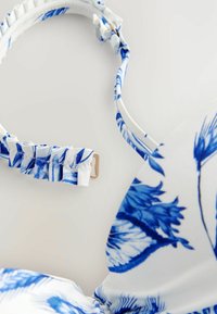 Top bikini bianco con stampa floreale blu, caratterizzato da una tracolla arricciata, una texture liscia e un fermo in tono dorato per la chiusura.