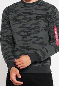 Felpa a motivo mimetico in verde scuro e nero. Caratterizzata da un collo tondo, polsini a coste e una patch sulla manica laterale con accento rosso.