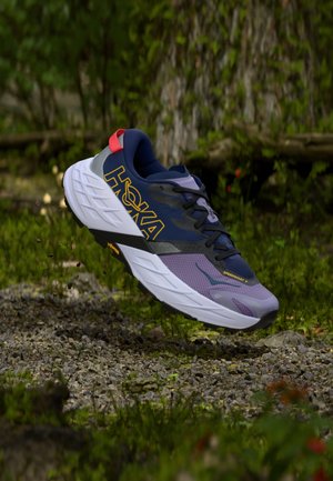 Pantof de alergare trail Hoka Speedgoat 7 în nuanțe de albastru închis și mov, suspendat deasupra unui traseu de pietriș în cadrul unei păduri verzi.