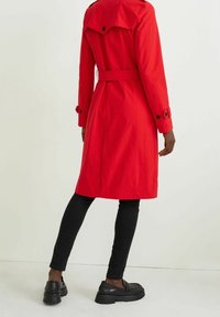 Trench rojo con cintura ceñida, detalles de botones en las mangas y una abertura trasera, combinado con pantalones negros y zapatos negros gruesos.