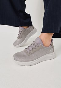 Persona que lleva zapatillas de malla gris claro con suelas blancas y pantalones azul marino a rayas finas, de pie sobre una superficie blanca.