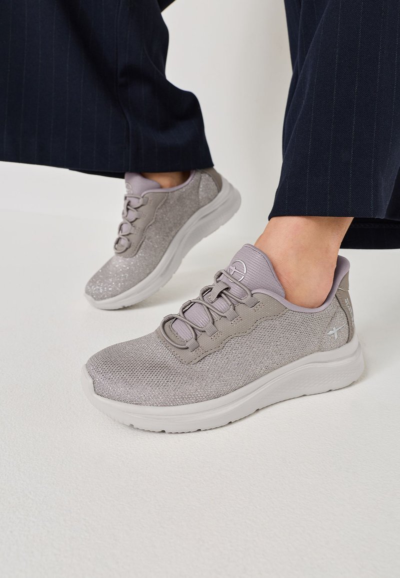 Persona que lleva zapatillas de malla gris claro con suelas blancas y pantalones azul marino a rayas finas, de pie sobre una superficie blanca.