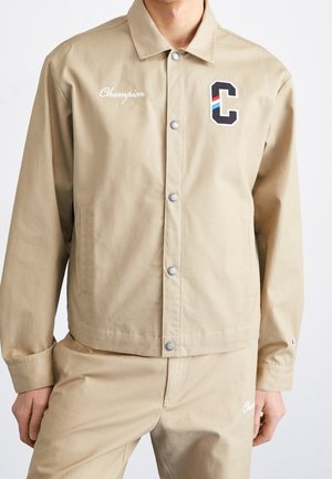 Homme portant une veste et un pantalon beige à boutons pression, avec "Champion" brodé sur la poitrine gauche et le pantalon, ainsi qu'un patch en forme de lettre "C" sur la poitrine droite.