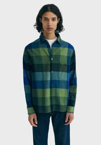 Persoon gekleed in een blauw en groen grote geruite button-up shirt over een wit onderhemd, gecombineerd met donkerblauwe jeans, staande tegen een eenvoudige achtergrond.