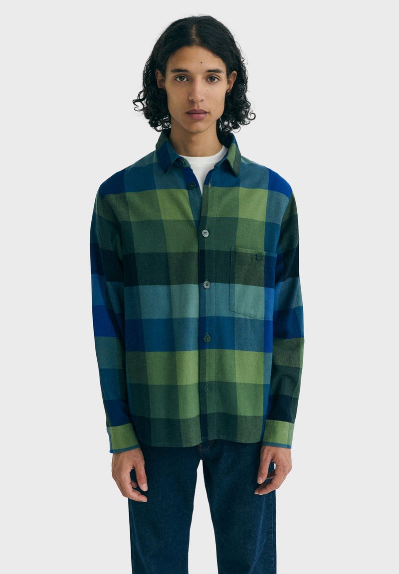 Persoon gekleed in een blauw en groen grote geruite button-up shirt over een wit onderhemd, gecombineerd met donkerblauwe jeans, staande tegen een eenvoudige achtergrond.
