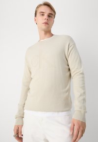 Maglione a maglia beige chiaro con logo "CK" in rilievo, polsini a coste e design classico a girocollo. Indossato sopra una camicia bianca.
