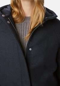 Giacca nera in misto lana con zipper, dettagli a scatto e cappuccio foderato, indossata sopra un maglione grigio a coste.