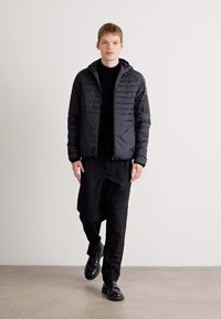 Jovem a andar dentro de casa, vestido com uma jaqueta preta acolchoada com capuz, calças pretas, sapatos pretos e uma camisa preta, em frente a uma parede lisa.