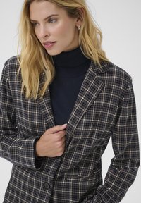 Marynarka w kratę w kolorze granatowym, beżowym i białym. Posiada klapy w stylu notched, zapięcie na jeden guzik oraz strukturalną tkaninę. Noszona na ciemnym golfie.