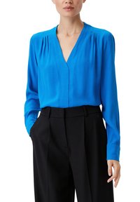 Blusa blu a scollo a V con maniche lunghe, caratterizzata da leggere pieghe sulle spalle, abbinata a pantaloni neri sartoriali. Tessuto liscio e leggero.