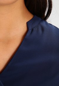 Blusa blu navy con una texture liscia, caratterizzata da un colletto a V e un leggero dettaglio sulle spalle, che mette in risalto la lucentezza del tessuto.