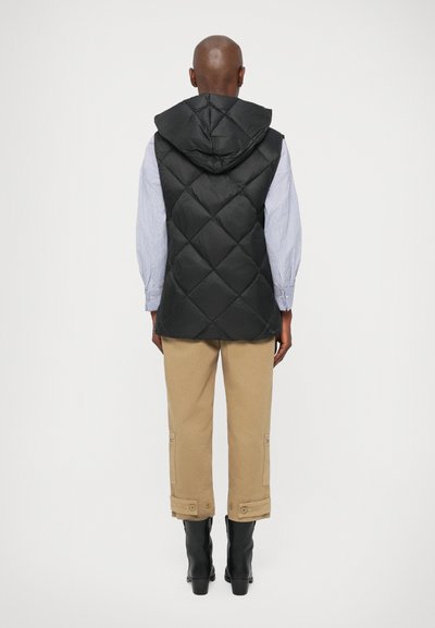 Must mustriga vest, millel on kapuuts ja millel on ruuduline muster. Kanti helesinise triibulise särk ja beežide pükste kohal, koos mustade saapadega.